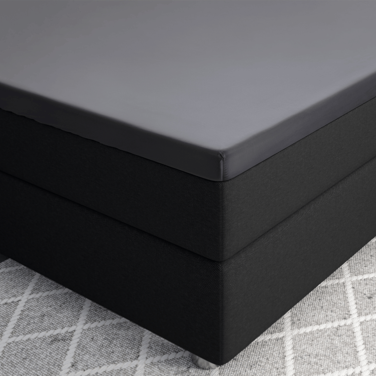 Nahaufnahme einer mit dem Primaviera Deluxe Topper Satin Deluxe Spannbettuch - Anthrazit bezogenen Matratzenecke auf einem dunklen Lattenrost, der auf einem hellen, gemusterten Teppich liegt.