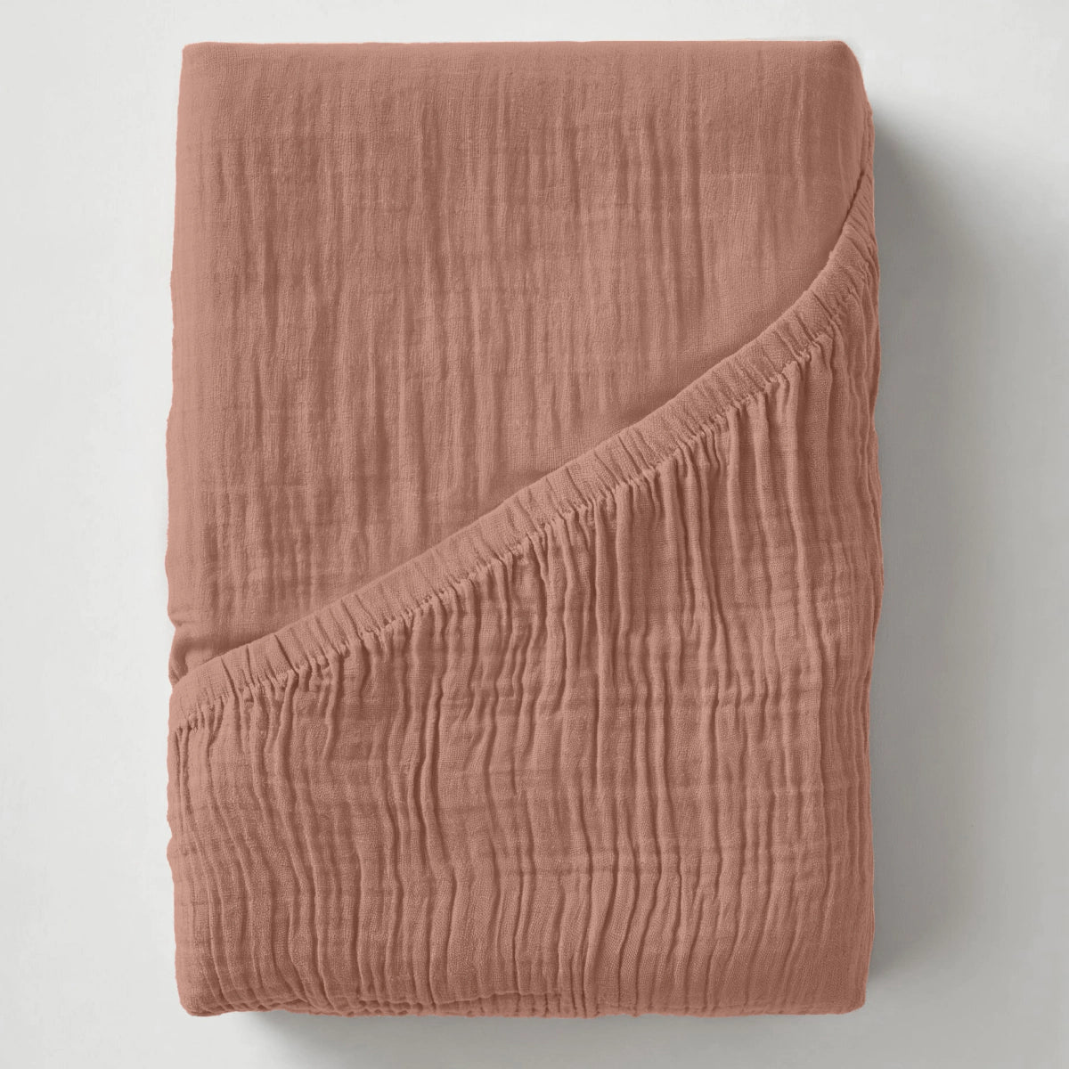 Ein gefaltetes, strukturiertes Musselin Spannbettlaken - Dusty-Rose mit einer umgeschlagenen Ecke wird auf einer schlichten weißen Fläche präsentiert.