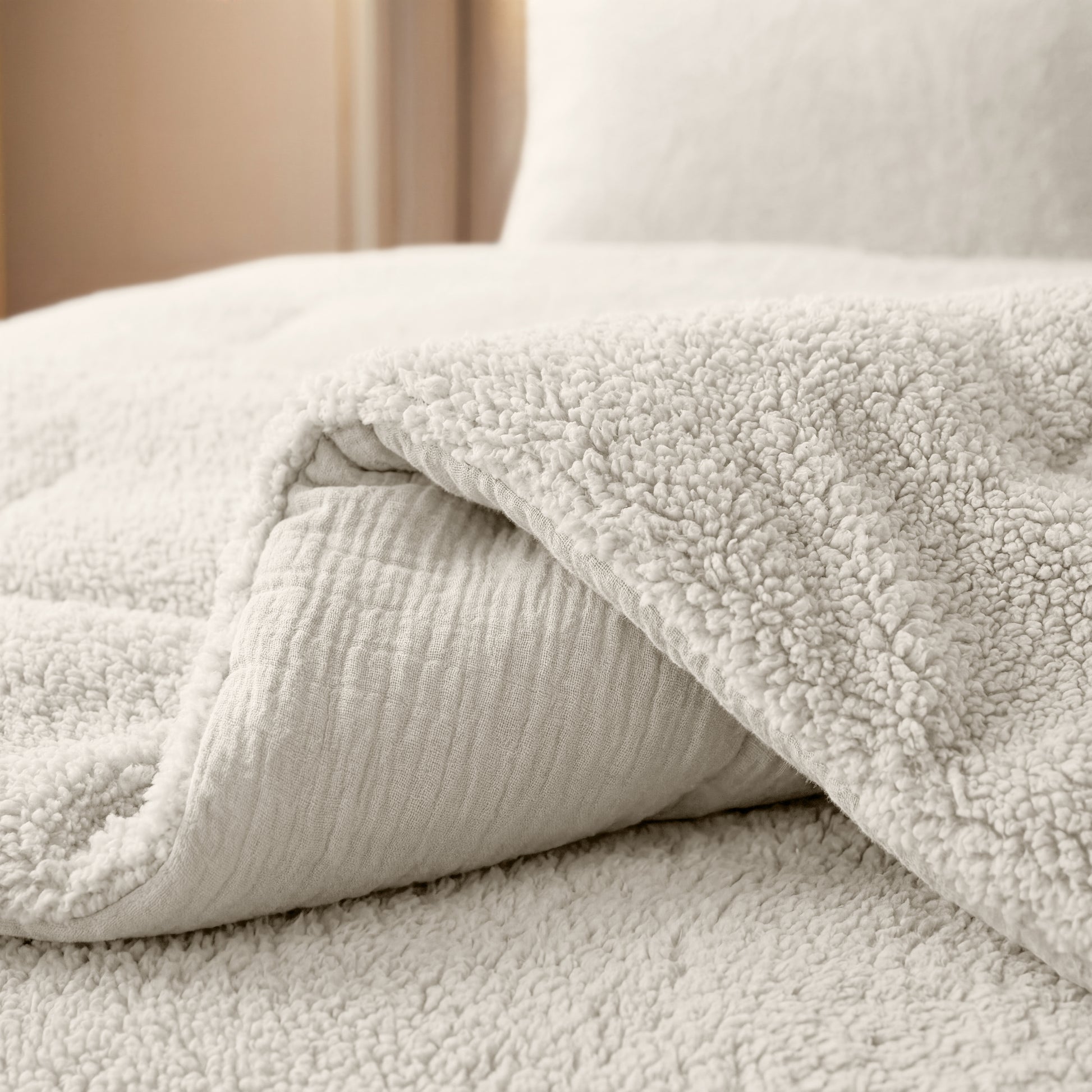 Eine Nahaufnahme eines ordentlich gemachten Bettes mit der Zelesta Teddy Baumwolle Musselin - Beige Bettdecke, deren strukturierte, cremefarbene Oberfläche teilweise zurückgeschlagen ist, um ein gestepptes Laken darunter freizulegen.