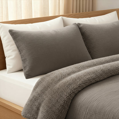 Ein ordentlich gemachtes Bett mit weißen Kissen, grauen strukturierten Kissenbezügen und einer passenden grauen Zelesta Teddy Baumwolle Musselin - Taupe Bettdecke in einem hölzernen Bettgestell neben beigen Vorhängen.