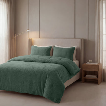 Ein ordentlich gemachtes Bett mit grüner Zelesta-Teddy-Baumwoll-Musselin-Bettwäsche und zwei Kissen steht vor einer beigen Wand, begleitet von einem Nachttisch und einer Stehlampe. Helle Vorhänge hängen über hohen Fenstern.