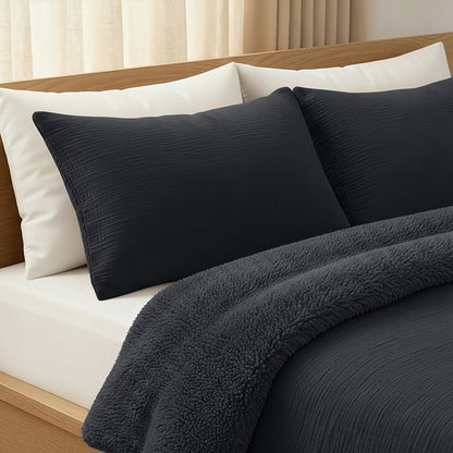 Ein ordentlich gemachtes Bett mit einem hellen Holzrahmen, weißen Kissen und der Zelesta Teddy Baumwolle Musselin - Anthrazit Bettdecke mit temperaturregulierender Füllung, umrahmt von cremefarbenen Vorhängen für eine gemütliche, einladende Atmosphäre.