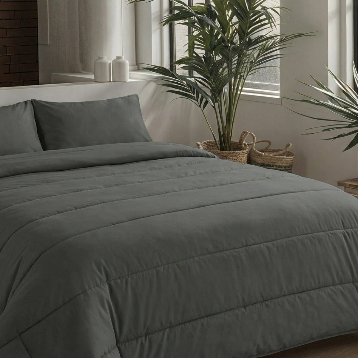 Ein ordentlich gemachtes Bett mit dunkelgrauer Mikrofaser-Bettwäsche und Zelesta Comfort Uni Kissenbezügen Set von 2 - Anthrazit (80x80cm) steht neben großen Fenstern und zwei Topfpflanzen in einem modernen, minimalistischen Schlafzimmer.