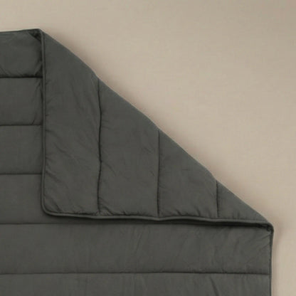 Eine Zelesta Comfort Uni Bettdecke in Anthrazit liegt flach auf einer beigen Oberfläche, wobei eine Ecke umgeschlagen ist, um die detaillierten Nähte und die weiche Textur zu zeigen.