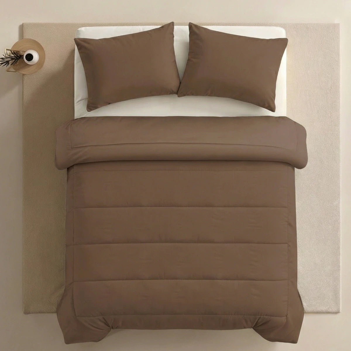 Ein ordentlich gemachtes Bett mit Zelesta Comfort Uni Kissenbezügen Set von 2 - Mokka (80x80cm) und einer passenden Bettdecke, aufgestellt auf einem hellbeigen Teppich neben einem kleinen runden Tisch mit einer Vase in der Ecke.