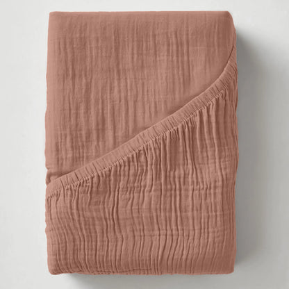 Ein gefaltetes, strukturiertes Musselin Spannbettlaken - Dusty-Rose mit einer umgeschlagenen Ecke wird auf einer schlichten weißen Fläche präsentiert.