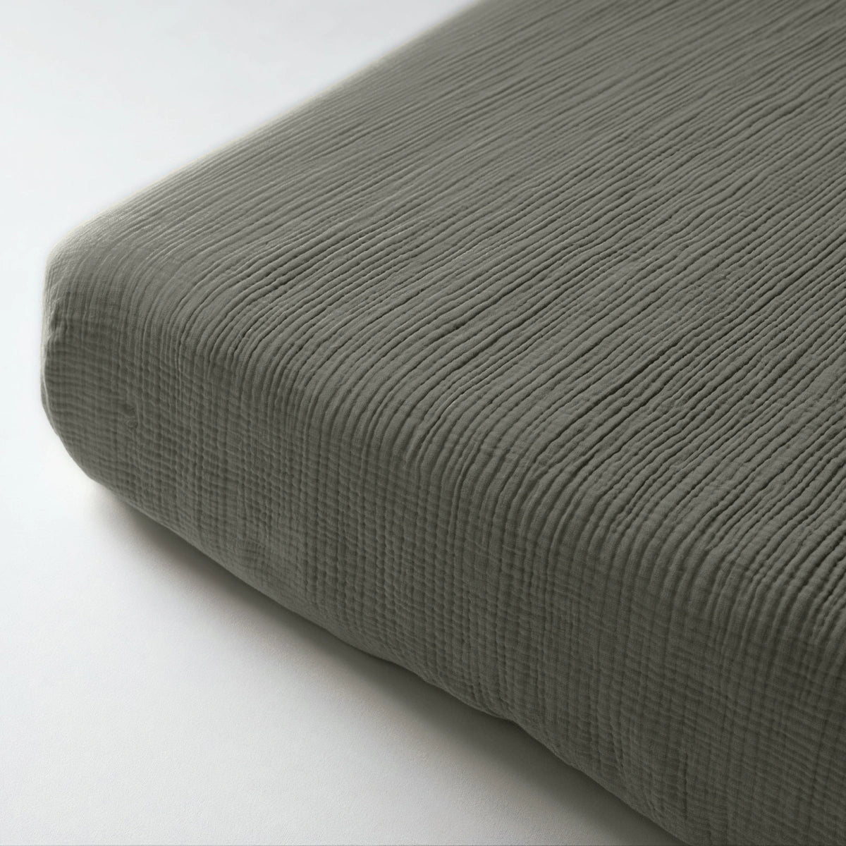 Nahaufnahme eines strukturierten Musselin-Spannbettlakens in Grau-Grün auf einer Matratze, wobei der gerippte Stoff und die minimalistische Eleganz vor einem schlichten Hintergrund hervorgehoben werden.