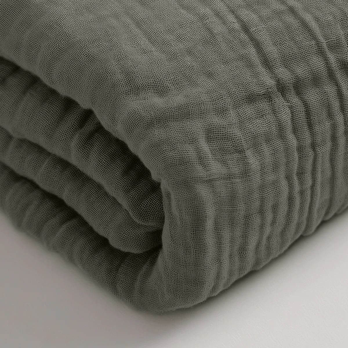 Eine Nahaufnahme eines gefalteten, strukturierten Musselin-Spannbettlakens - Grau-Grün auf einer weißen Fläche.