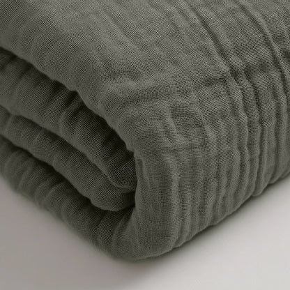 Eine Nahaufnahme eines gefalteten, strukturierten Musselin-Spannbettlakens - Grau-Grün auf einer weißen Fläche.