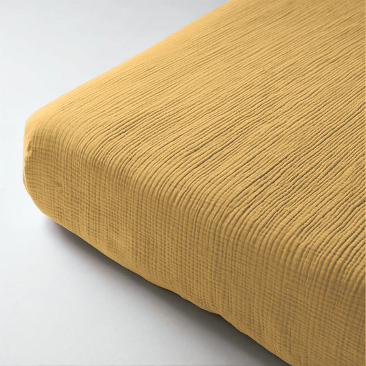Eine Nahaufnahme des Musselin-Spannbettlakens in Ockergelb mit seiner gerippten Struktur, die eine Matratzenecke auf einer hellen Fläche bedeckt.
