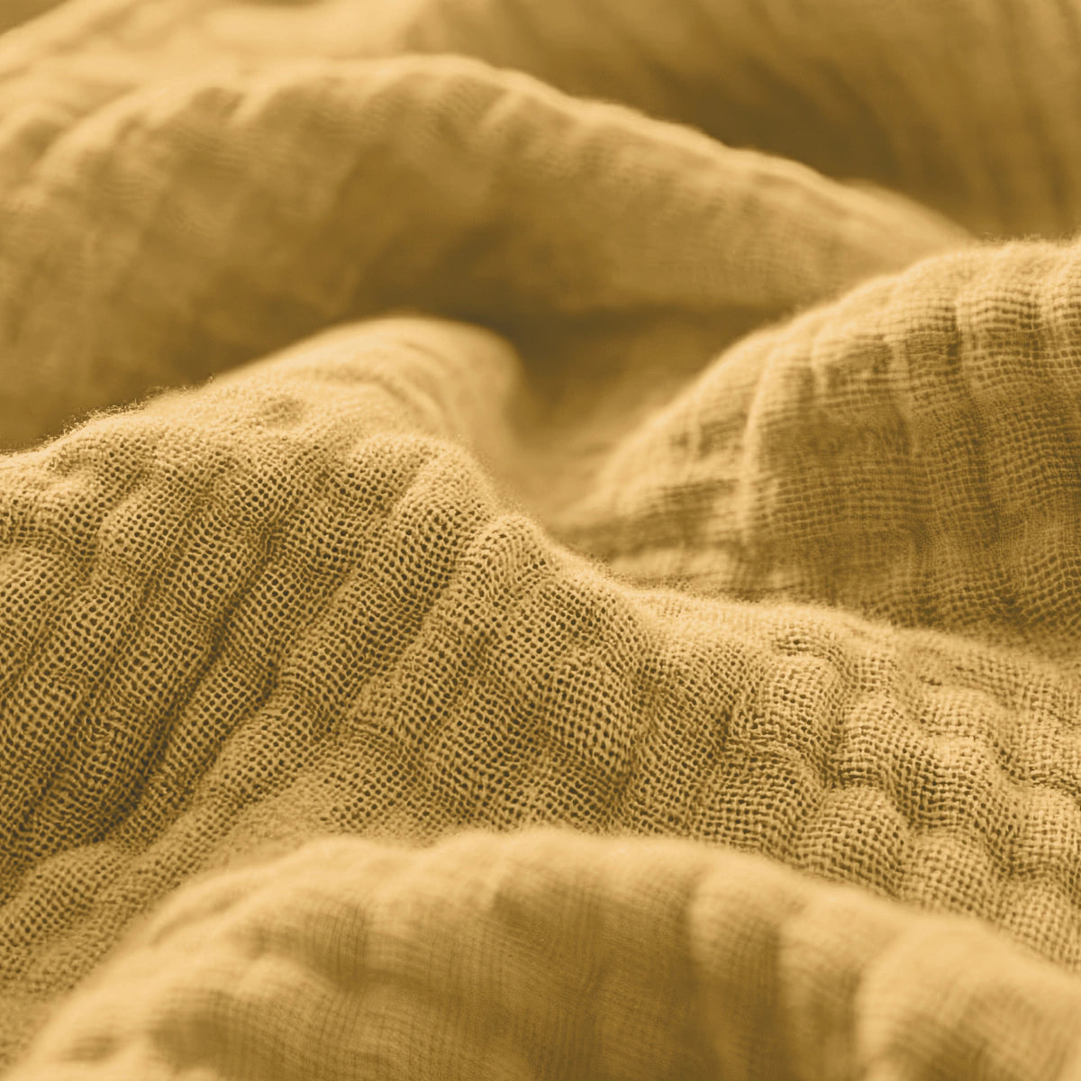 Nahaufnahme des strukturierten, faltigen Musselin-Spannbettlakens in Ockergelb, das seine gewebte Struktur und die weichen Falten betont.