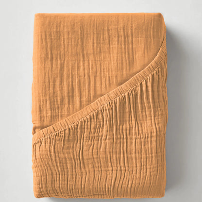 Ein gefaltetes Musselin Spannbettlaken - Taupe mit einer strukturierten Oberfläche und einer umgedrehten Ecke, dargestellt auf einem unifarbenen Hintergrund.