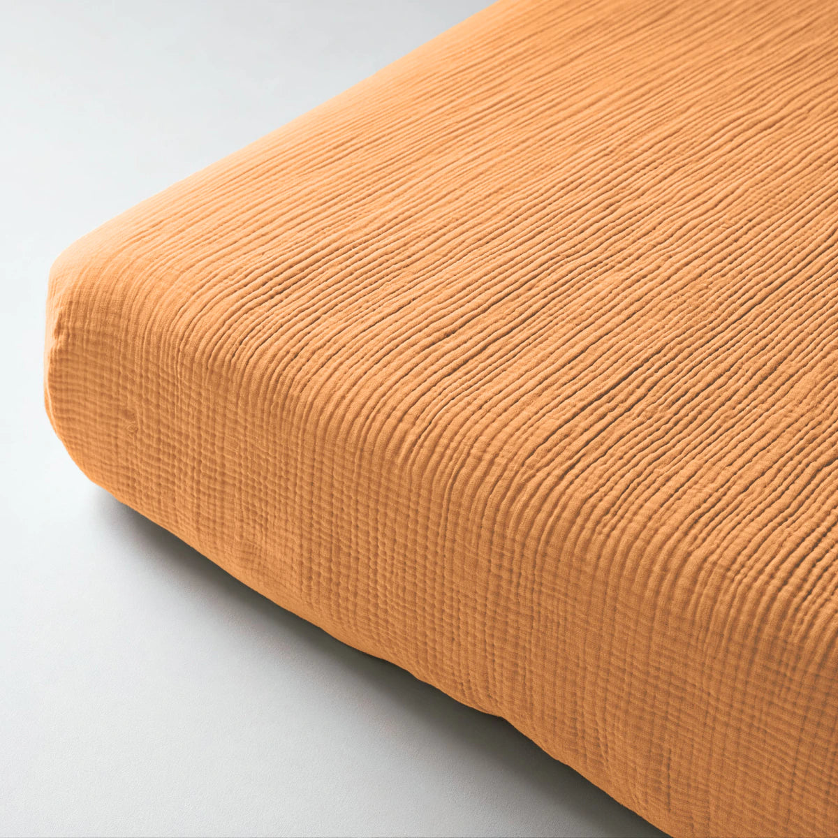 Nahaufnahme eines Musselin Spannbettlakens in Taupe, das eine Matratze bedeckt, vor einem schlichten hellen Hintergrund.