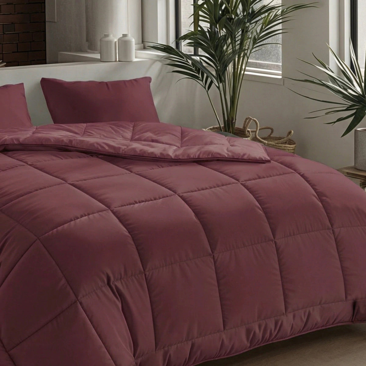 Ein ordentlich gemachtes Bett mit der Zelesta Pure Uni Bettdecke in Bordeauxrot und passenden Kissen steht neben einem Fenster, das mit Grünpflanzen und dekorativen Vasen auf einem Beistelltisch dekoriert ist.