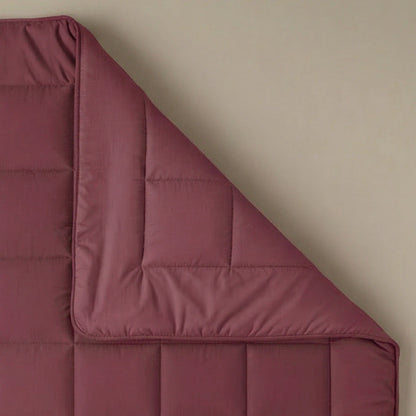 Die Zelesta Pure Uni Bettdecke in Bordeauxrot liegt fein säuberlich ausgebreitet auf einer beigen Unterlage, eine Ecke ist umgeschlagen und gibt den Blick auf die detaillierte Steppung frei.