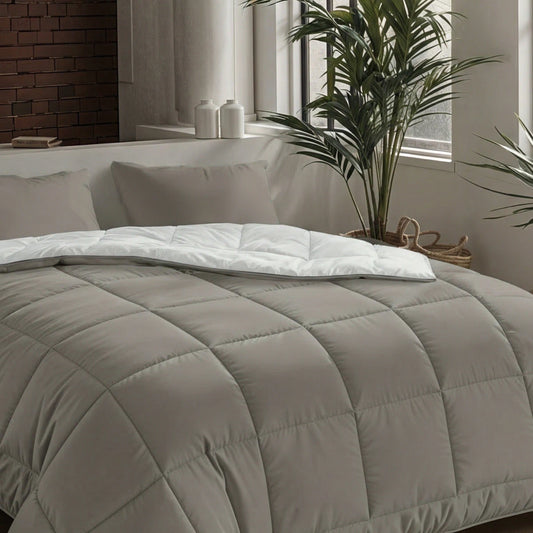 Ein ordentlich gemachtes Bett mit der Zelesta Pure Uni Bettdecke in Anthrazitgrau, weißen Laken und grauen Kissen neben einem Fenster mit Topfpflanzen und zwei weißen Deko-Gläsern auf der Fensterbank.